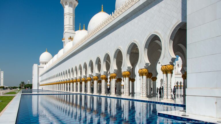 Long white arcade and gold-topped columns reflected in a blue courtyard pool.