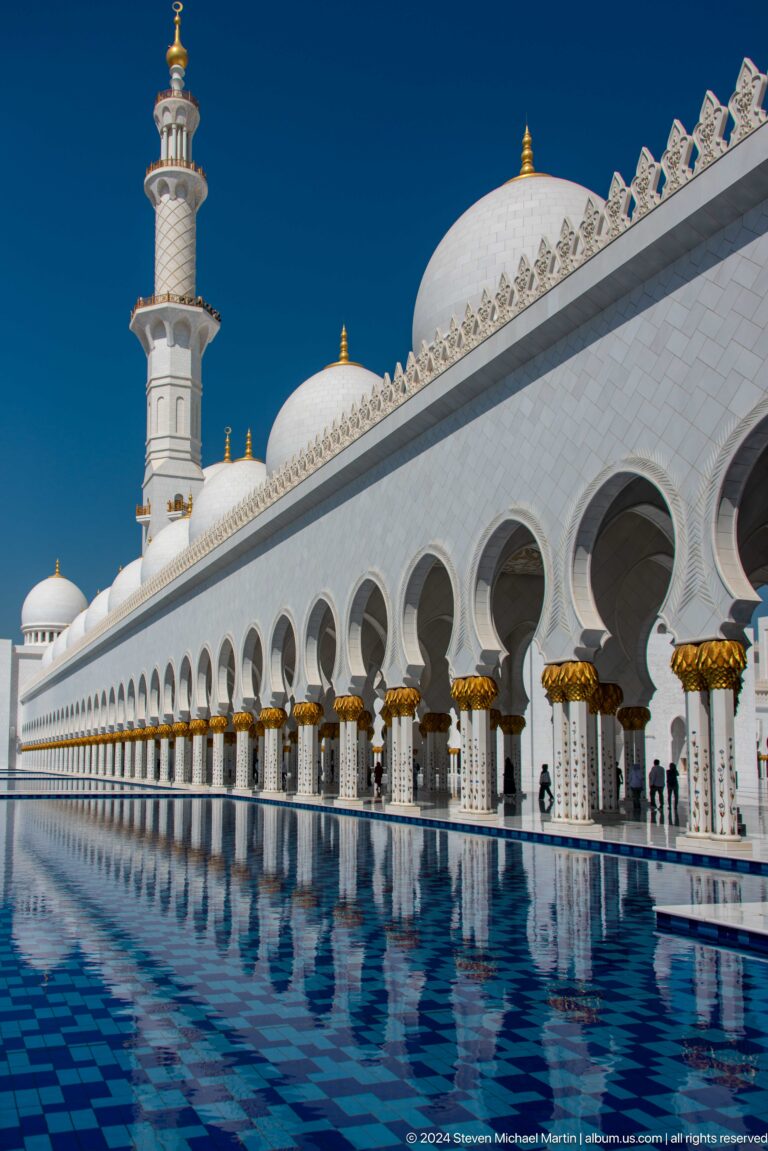 Visitors walk beneath a long white arcade beside a blue reflecting pool.