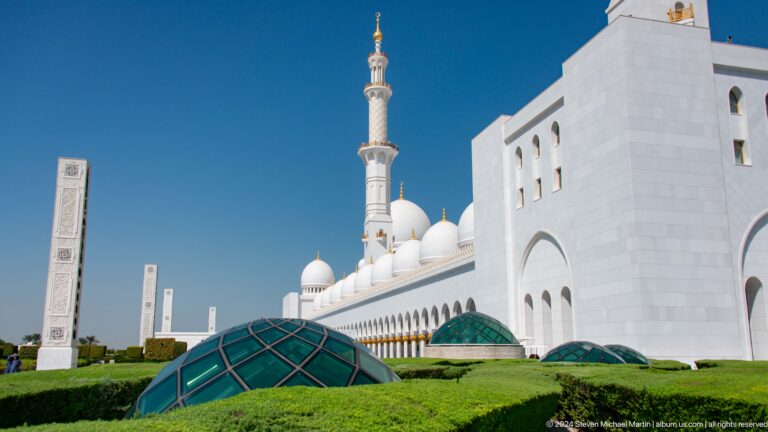 White mosque side elevation with minaret, domes, landscaping, and green covered structures.