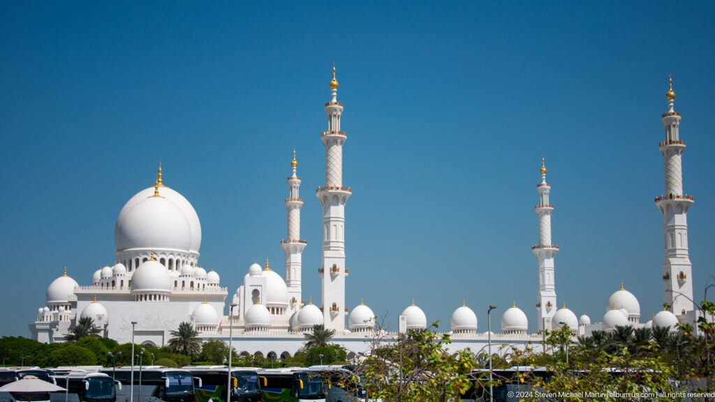 Wide view of white domes and four minarets rising above trees under a blue sky.