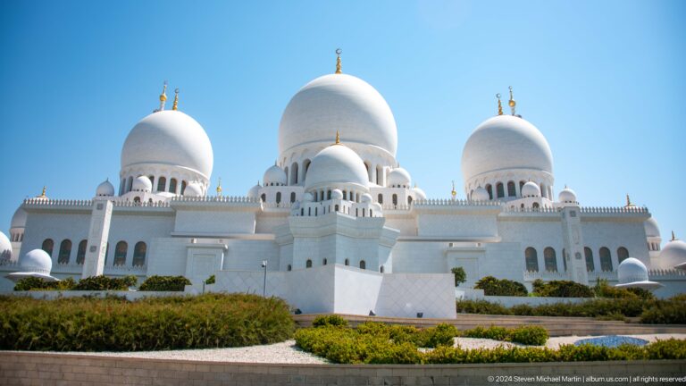 Three large white domes and smaller cupolas rise above an arched façade and landscaped beds.