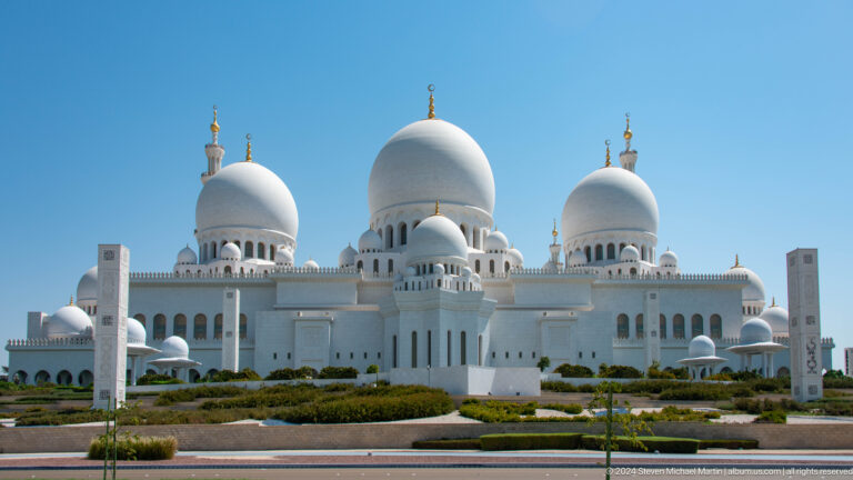 Frontal exterior view of three major white domes above a long arched façade.