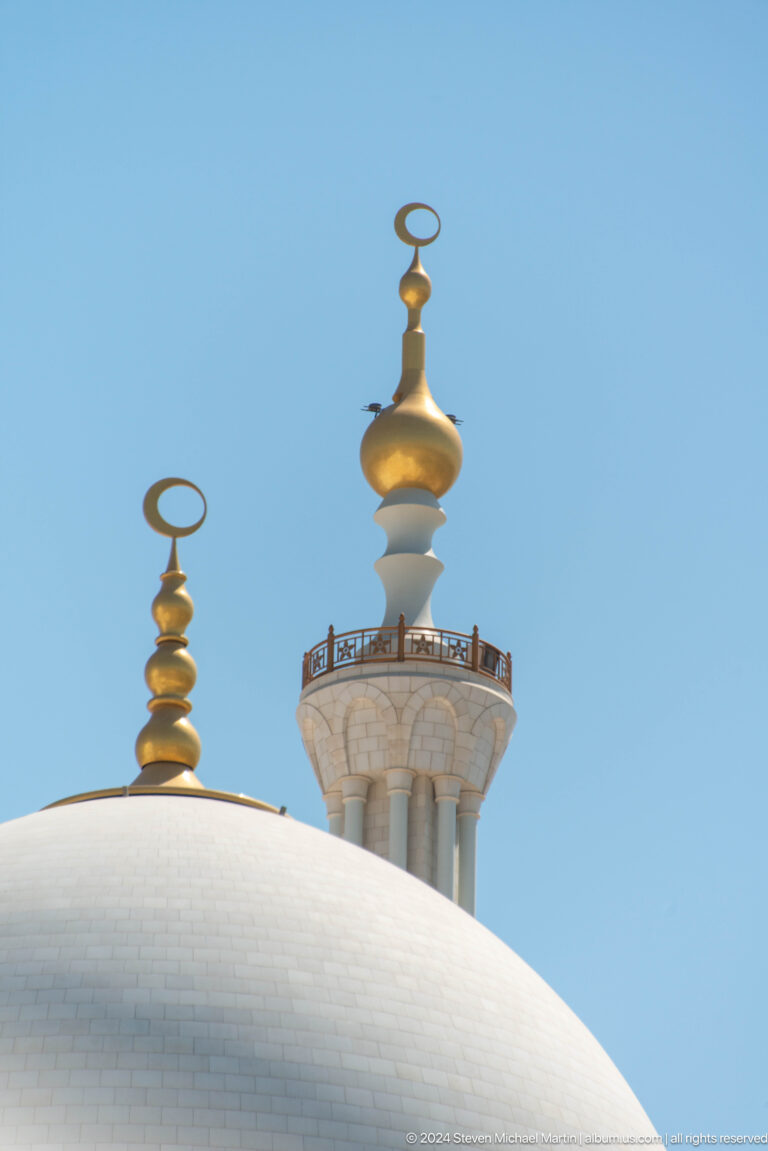 Gold crescent and stacked finial ornaments crowning a white minaret against blue sky.