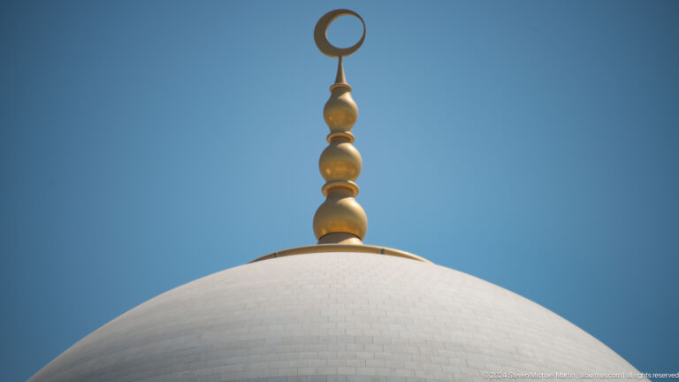 Gold crescent finial above a white mosque dome against a clear blue sky.