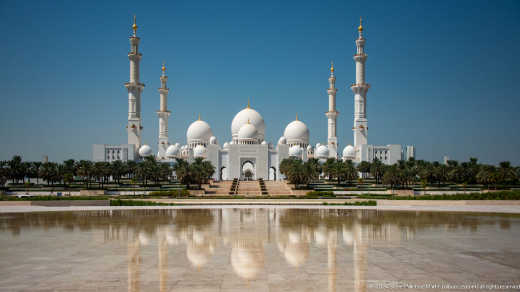 Frontal view of Sheikh Zayed Grand Mosque with domes, four minarets, palm trees, and a broad reflection.