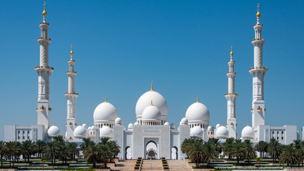 Vertical frontal view of the mosque's central entrance, white domes, four minarets, and date palms.