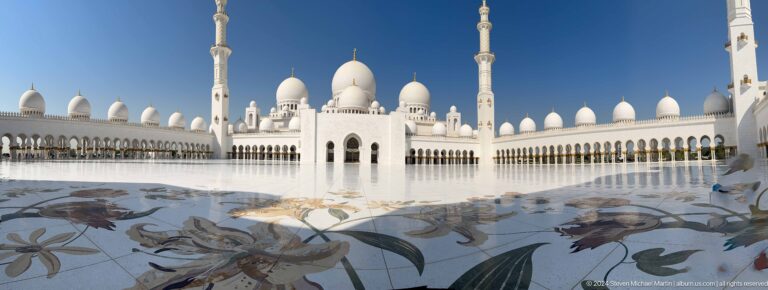 Large floral marble inlay in the mosque courtyard with white domes, arcades, and minarets beyond.