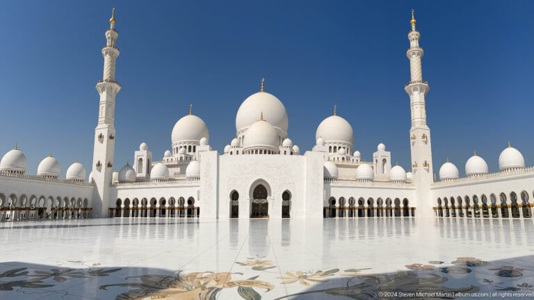 Centered courtyard view of the mosque's domes, twin minarets, arcades, and polished marble pavement.