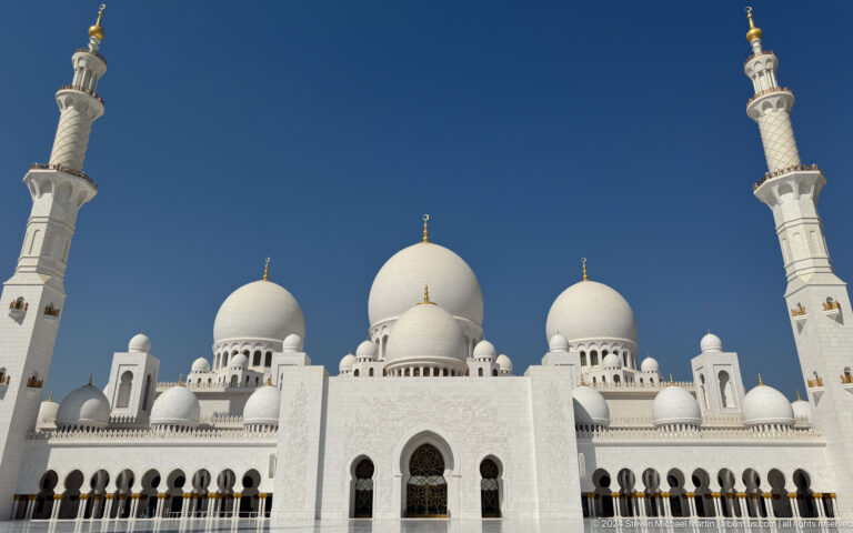 Low-angle view of clustered white domes and two minarets against a deep blue sky.