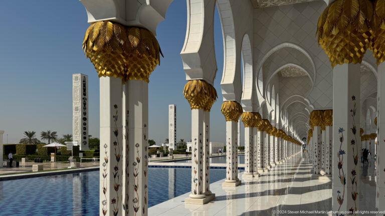 Sunlit white colonnade with gold palm capitals and floral columns beside a blue reflecting pool.