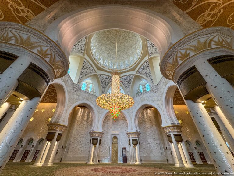 Wide interior view of massive white columns, a decorated dome, central chandelier, arches, and green carpet.