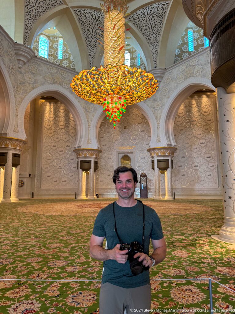 Male visitor standing beneath a gold chandelier in the mosque's ornate main prayer hall.