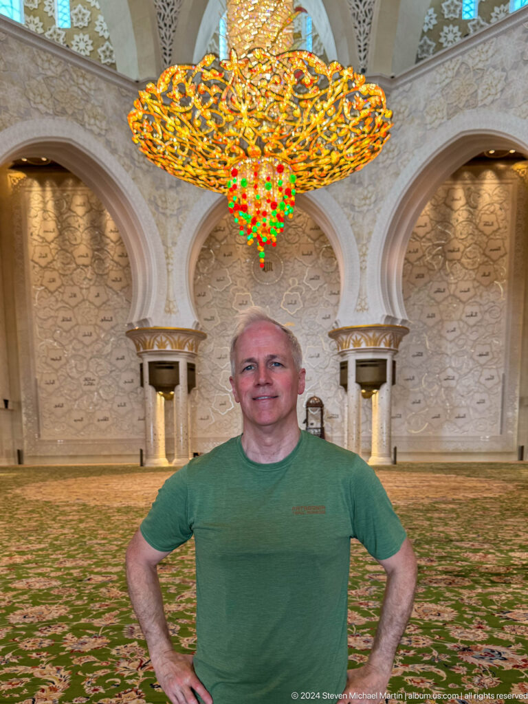 Gray-haired male visitor in a green shirt before the chandelier and decorated qibla wall.