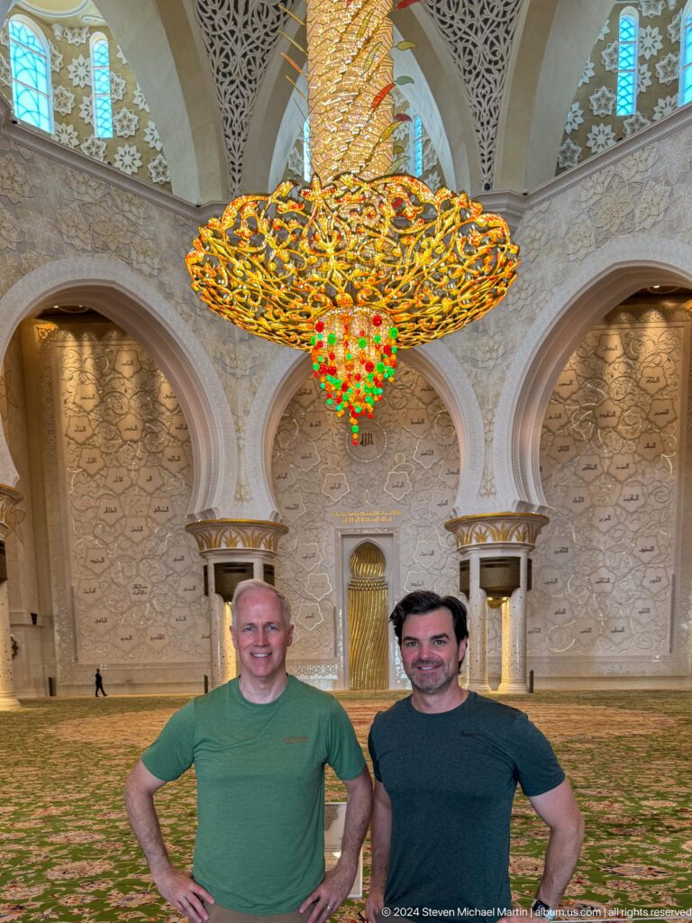 Two male visitors beneath a large gold chandelier in front of the mosque's decorated qibla wall.