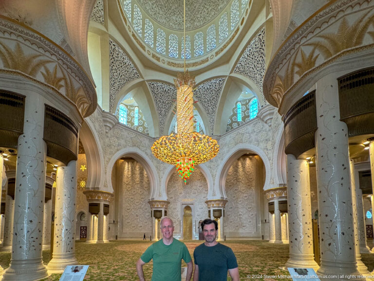 Two male visitors beneath a large chandelier inside a marble prayer hall with arches, columns, and floral carpet.