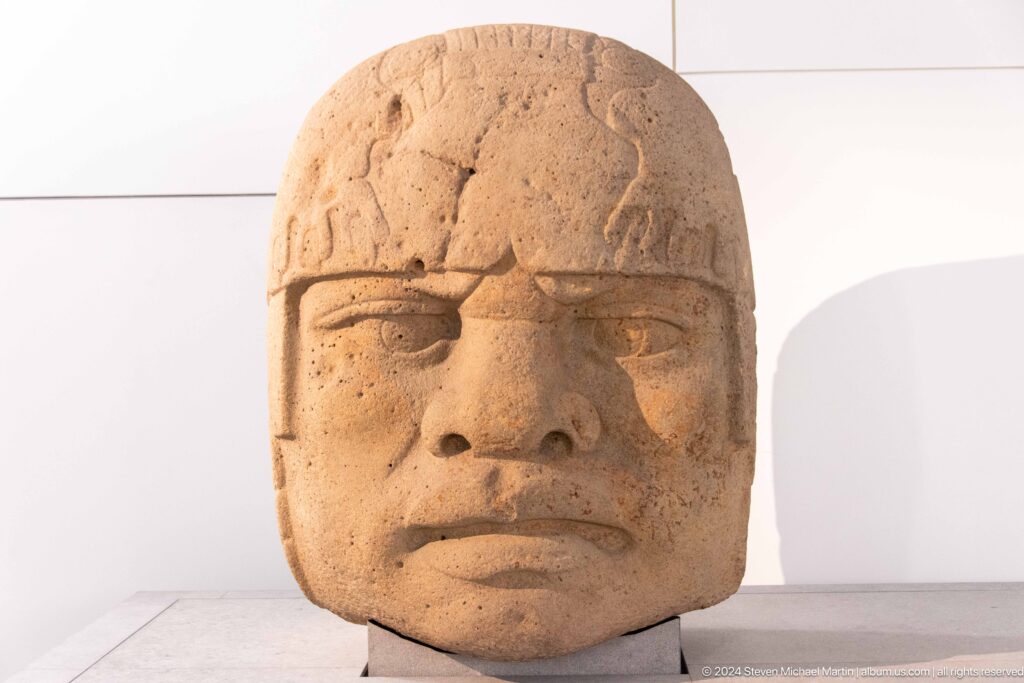 Large carved stone head on white plinth at Louvre Abu Dhabi.