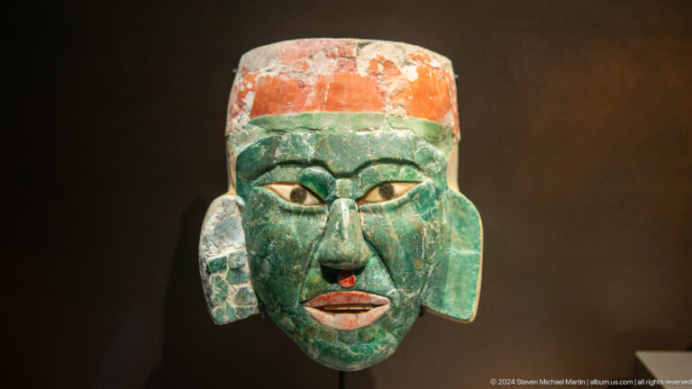 Green stone mask with red and white details at Louvre Abu Dhabi.