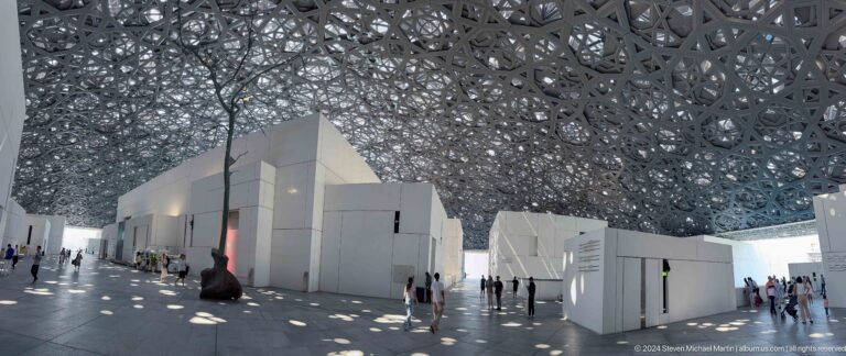 White gallery buildings beneath patterned dome at Louvre Abu Dhabi.