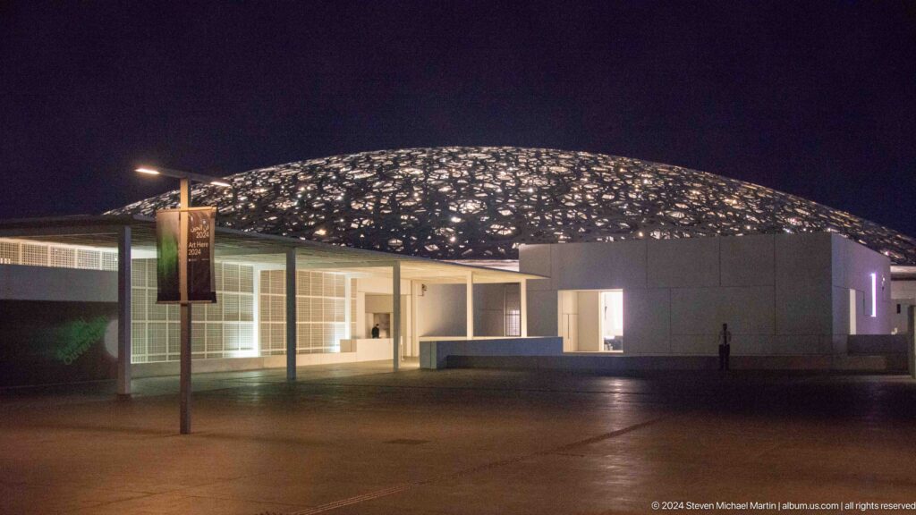 Museum dome glowing above low buildings at night at Louvre Abu Dhabi.