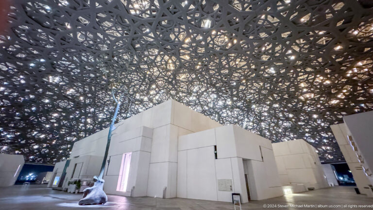 White gallery block under dense dome pattern at Louvre Abu Dhabi.