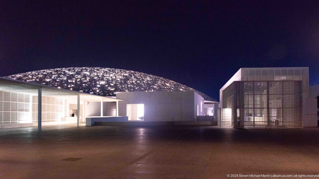 Low white galleries beneath illuminated dome at Louvre Abu Dhabi.