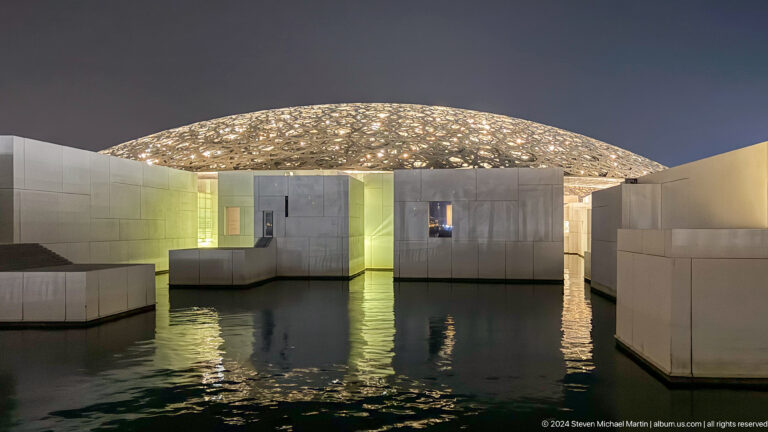 Illuminated dome and galleries reflected in water at Louvre Abu Dhabi.