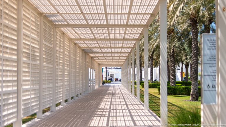 Sunlit lattice-covered walkway with palms at Louvre Abu Dhabi.