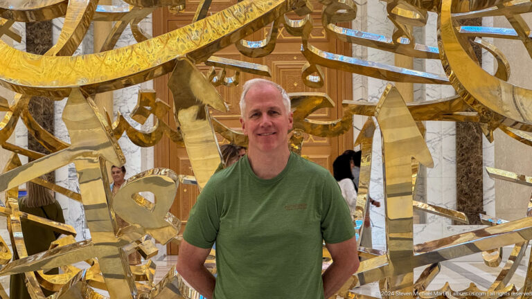 Visitor in green shirt before mirrored gold sculpture.