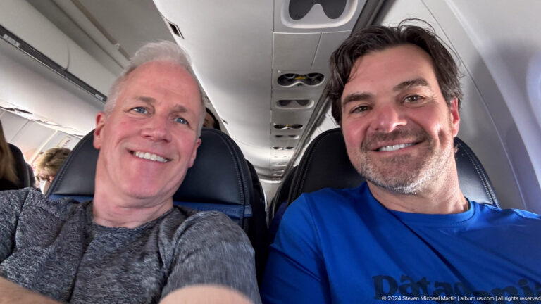 Two smiling men seated together inside an airplane cabin.