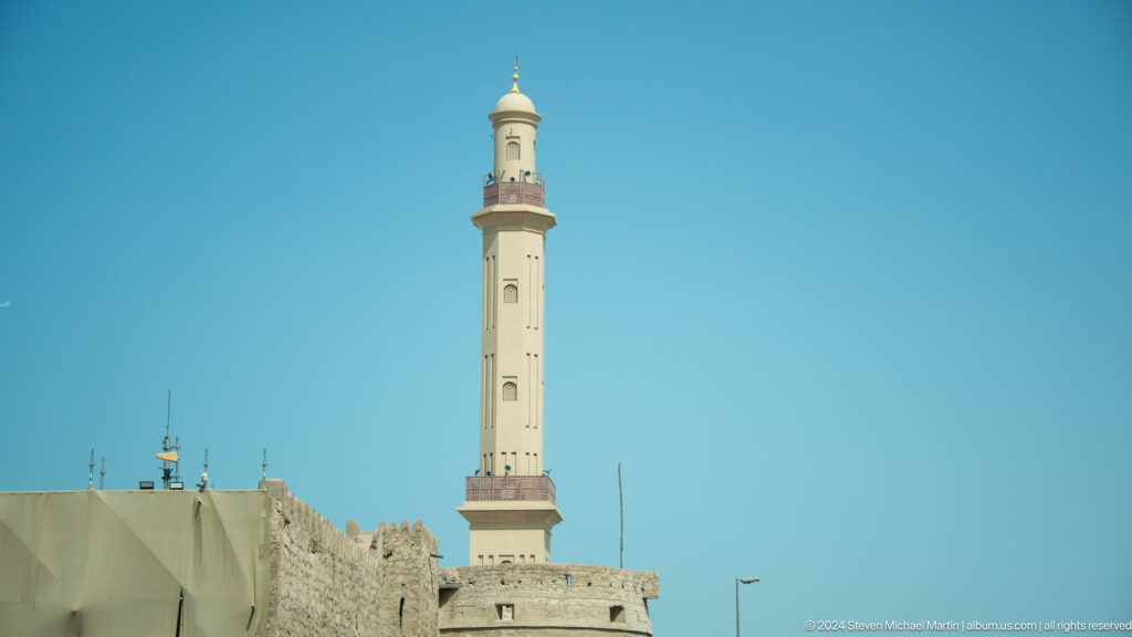 Tall beige minaret above low historic buildings in Dubai.