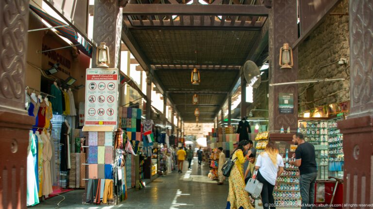 Covered market passage lined with textiles, shops, and shoppers.