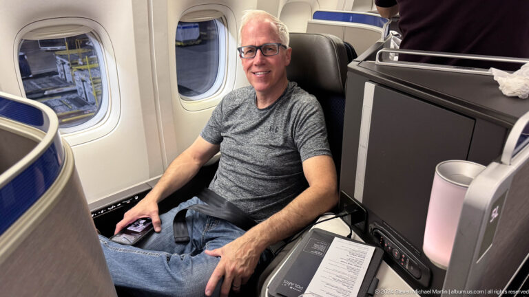 Smiling man in a gray shirt seated in a spacious airplane seat.