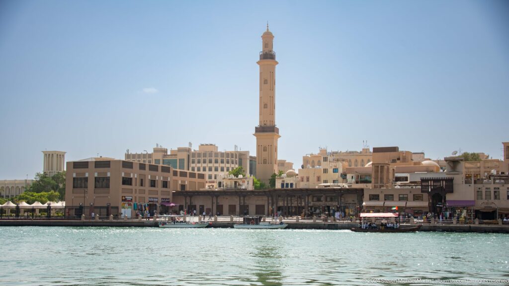 Tall mosque minaret centered above Dubai Creek waterfront buildings.