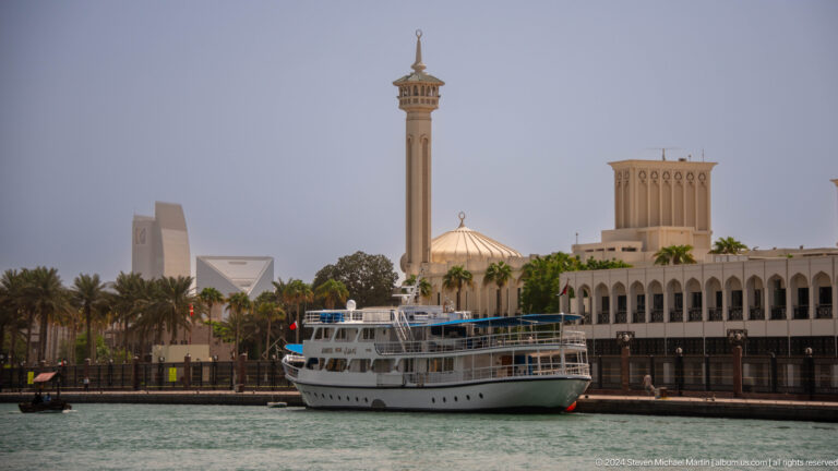 White passenger boat on Dubai Creek before wind towers and a minaret.