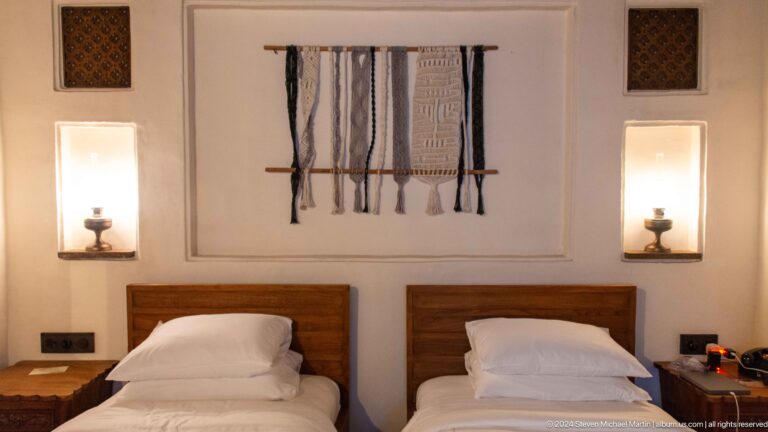 Heritage-style hotel room with two neatly made twin beds, dark wood headboards, woven textile artwork between them, and matching wall lamps.