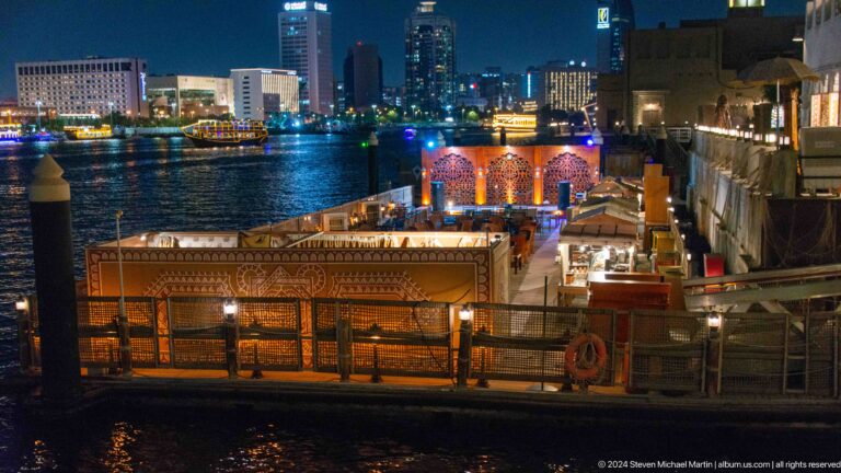 Night view of the Al Seef waterfront promenade with warmly lit, low-rise heritage-style buildings along Dubai Creek and small docks extending into the water.