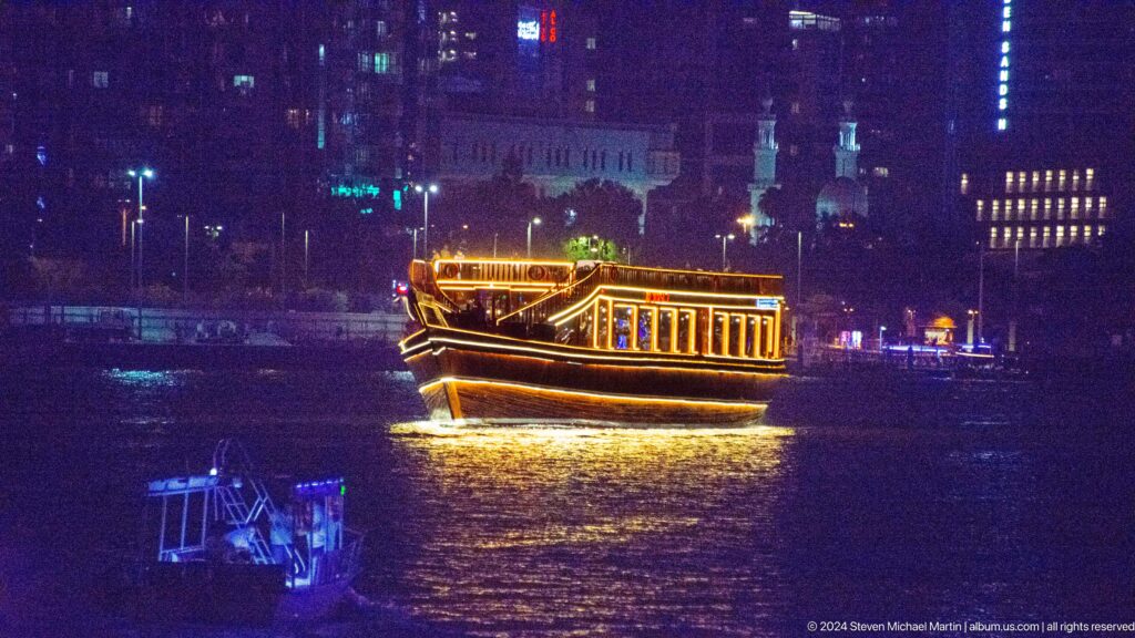 Multi-level tour boat outlined in warm lights cruising on dark water with a brightly lit city skyline behind.