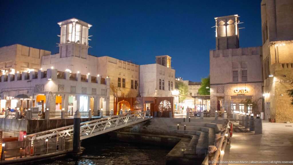 Night view of Al Seef waterfront with a low pedestrian bridge leading to cream-colored heritage-style buildings featuring a square wind tower and plastered walls.