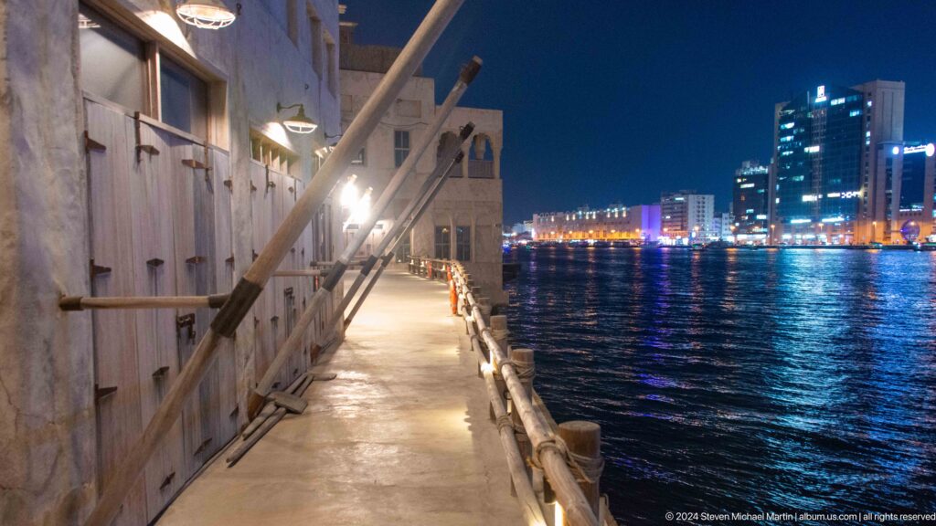Night view of a narrow creekside promenade at Al Seef along Dubai Creek, with a timber railing and wall-mounted lamps beside the dark water and the modern skyline beyond.