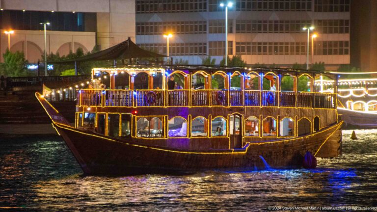 Nighttime view of a large two-deck cruise boat on Dubai Creek with dense lines of blue, yellow, and red decorative lights outlining its railings and windows.