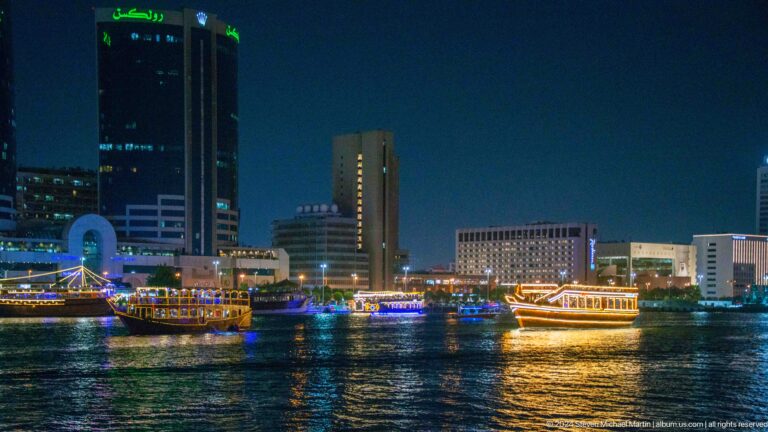 Night view of several illuminated boats crossing Dubai Creek with a continuous backdrop of modern office and hotel buildings.