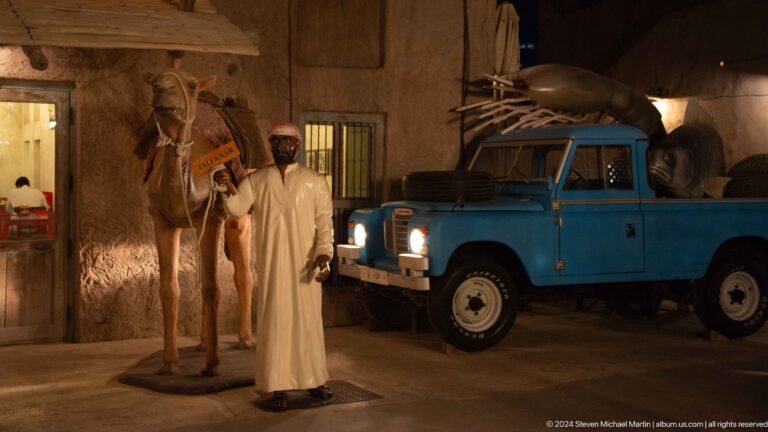 Man in traditional Gulf clothing standing beside a camel and a vintage blue utility vehicle in a nighttime heritage display at Al Seef Dubai.