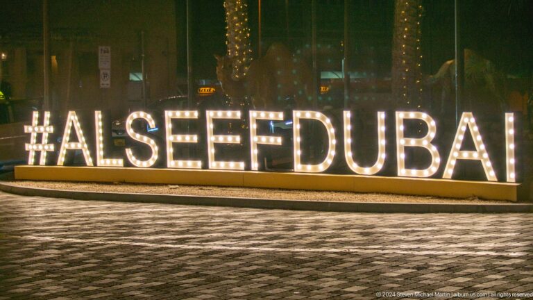 Nighttime view of a large illuminated AL SEEF DUBAI sign beside dark rippling waterfront.