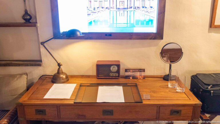 Wooden heritage-style writing desk in an Al Seef Dubai guest room, with writing materials, desk lamp, small mirror, and wall-mounted television.