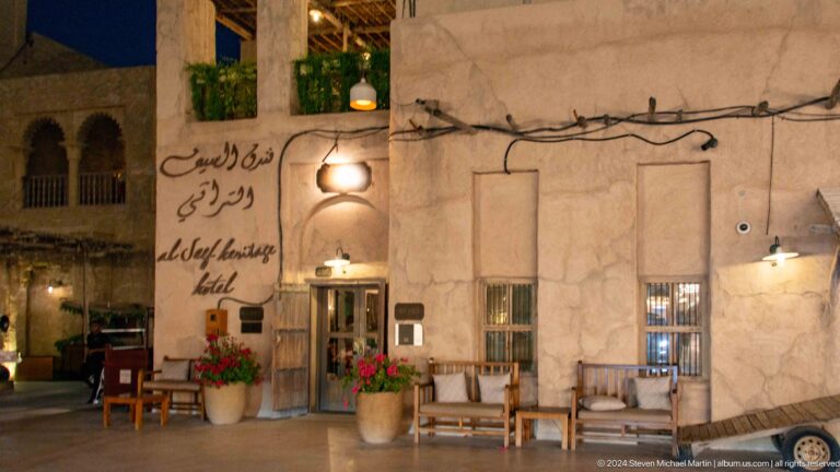 Outdoor café seating against a rough plaster façade with hanging lamps and handwritten-style wall text in Dubai’s Al Seef heritage district.
