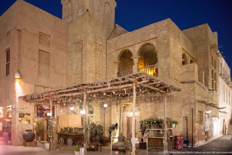 Night view of a two‑story heritage-style restaurant in Al Seef with arched upper openings and a wooden pergola sheltering a lit outdoor dining terrace.