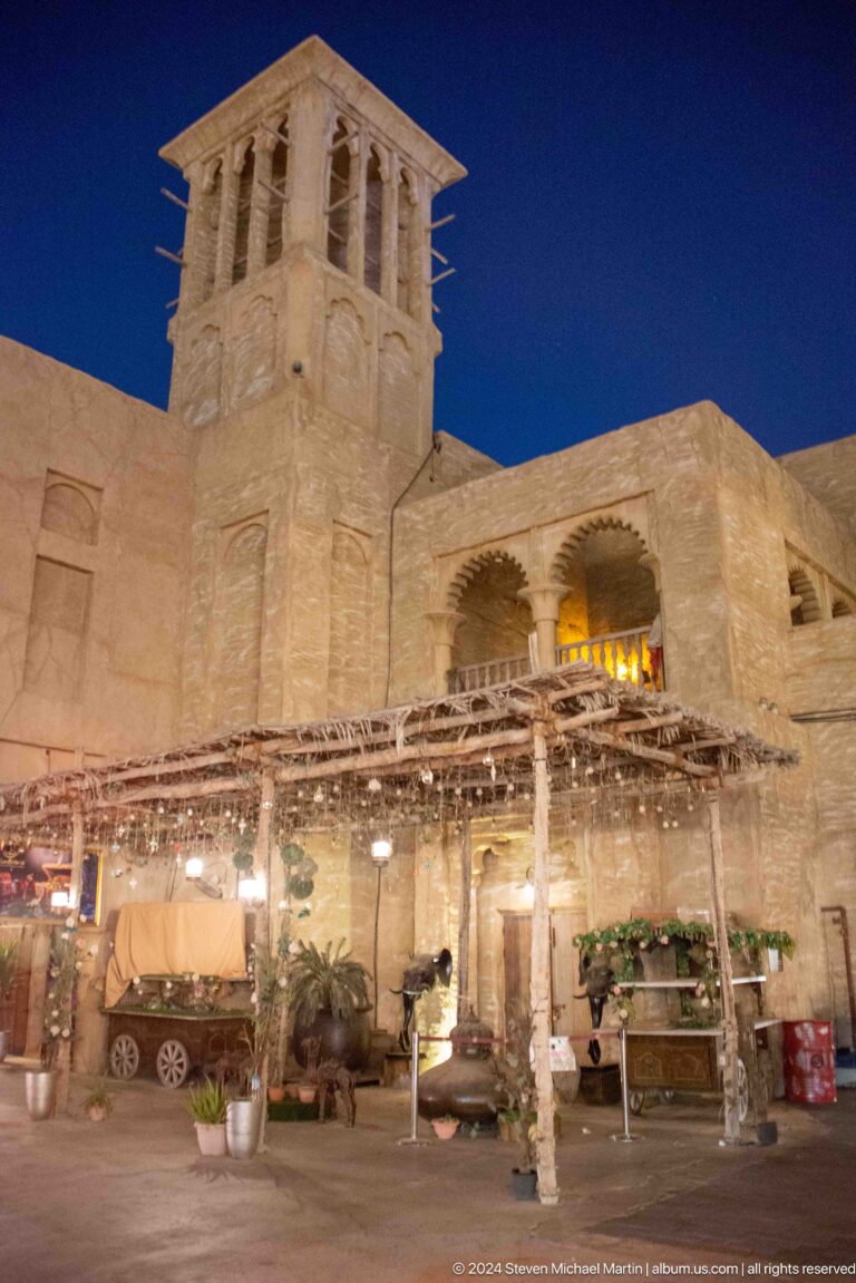 Detail of an Al Seef heritage-style restaurant façade with a deep arched upper opening above closely spaced timber pergola beams and rough plaster walls.