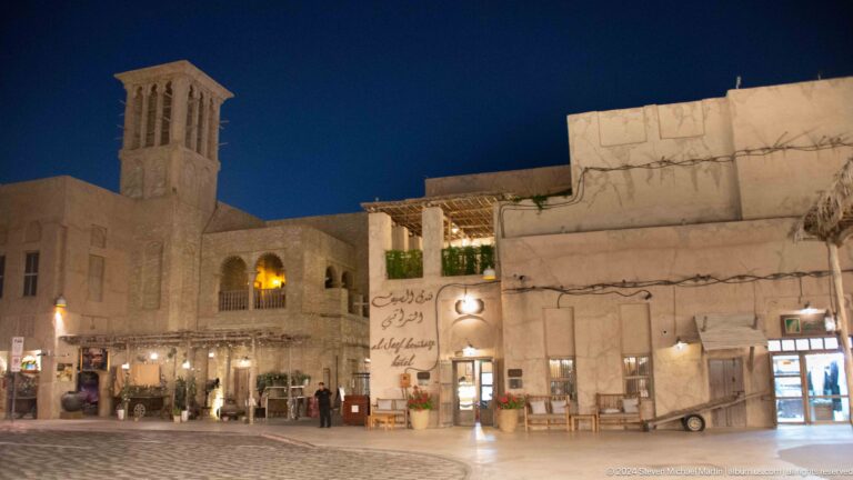 Night view of Al Seef streetscape with cream-colored heritage-style low-rise buildings and illuminated ground-floor shops along a pedestrian street.