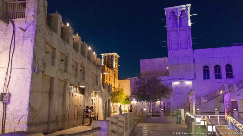 Night view of a narrow lane at Al Seef in Dubai with cream-colored rough plaster buildings and an illuminated square wind tower above the roofs.
