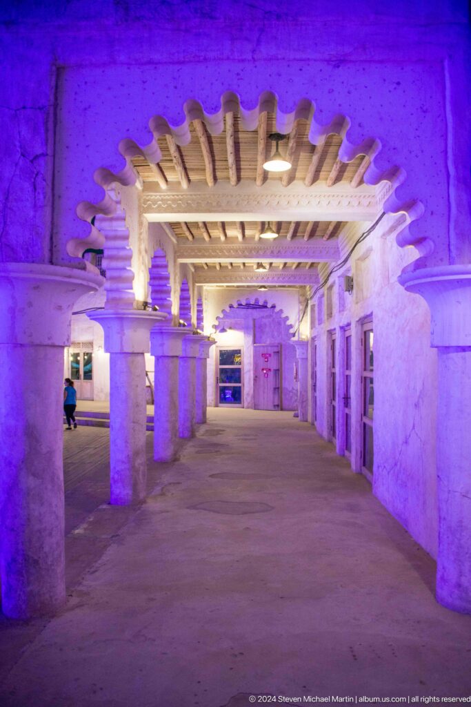 Long covered arcade in Al Seef, Dubai, with thick square columns, timber ceiling beams, scalloped arches, and purple lighting creating a strong sense of depth.